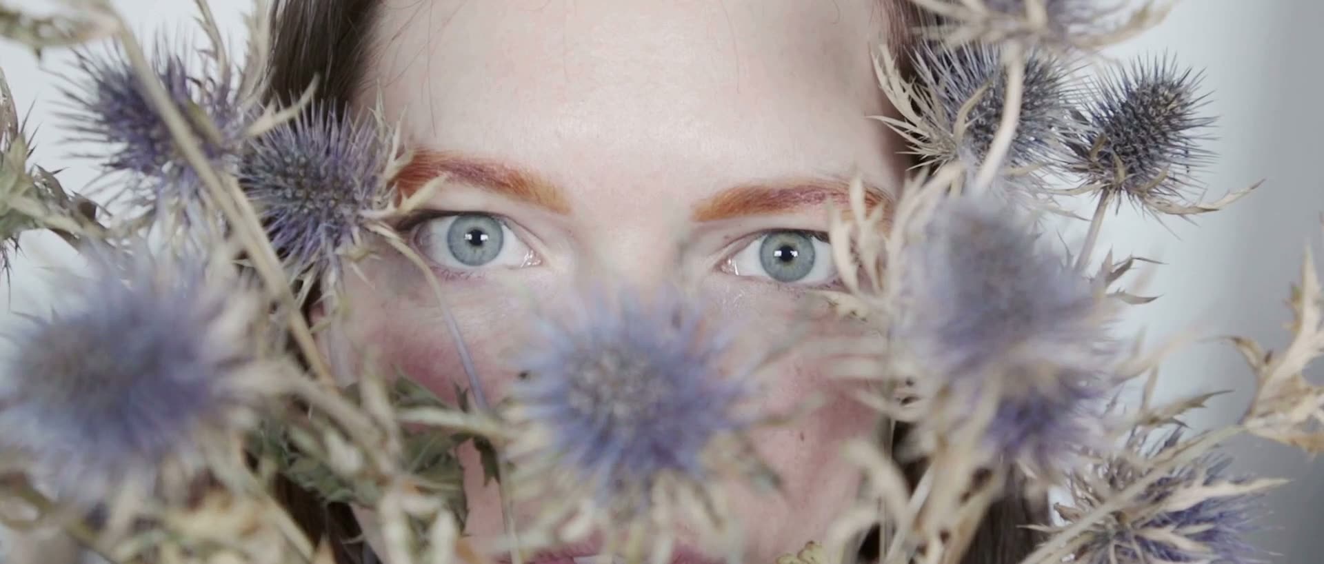 Close-up of a woman's blue eyes framed by dried thistle flowers, the opening frame of the Transform& brand film